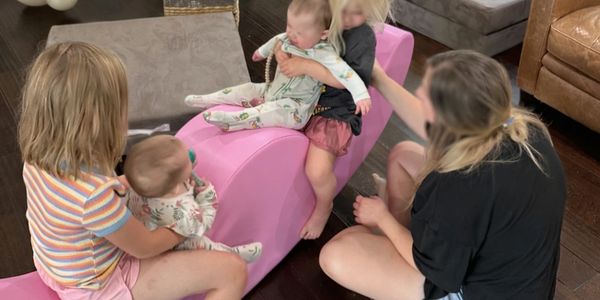 Children and a woman playing together indoors with a pink foam rocker.
