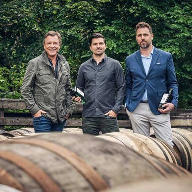 Three men holding gin bottles in front of wooden barrels in a natural setting—spirits experts