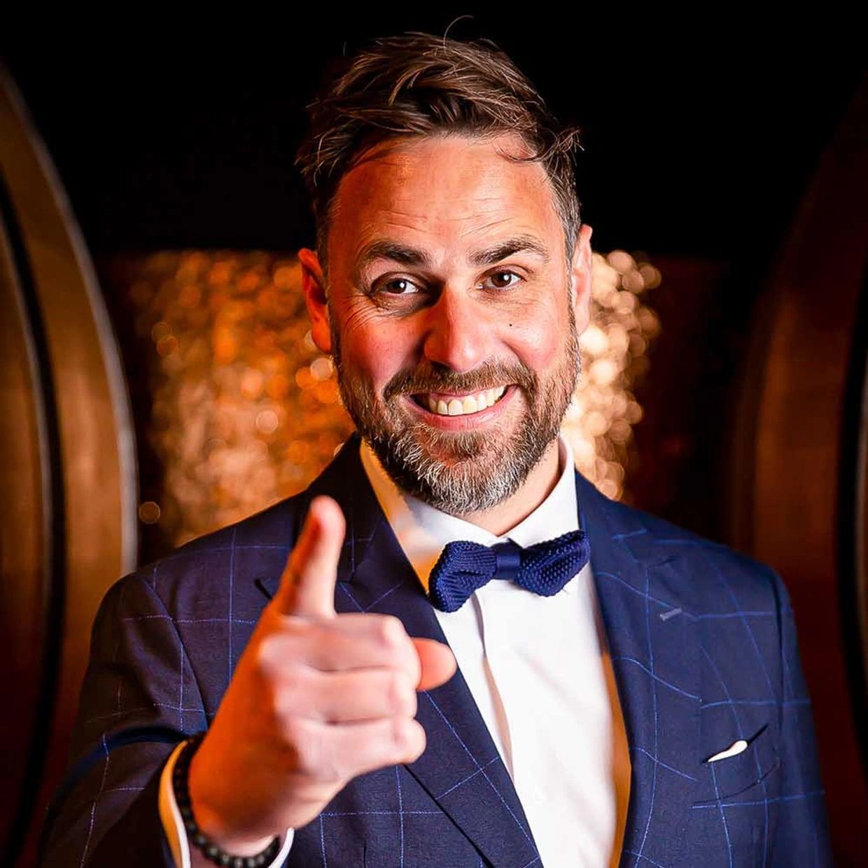 Peter Jauch im eleganten blauen Anzug mit Fliege, lächelnd und mit ausgestrecktem Finger zur Kamera – charismatischer Experte für Champagner, Gin und Genussmomente.