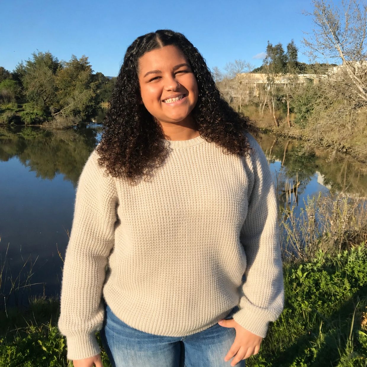 Smiling woman with curly hair standing by a lake on a sunny day.