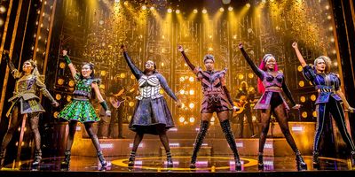 Six performers in vibrant costumes strike a triumphant pose on stage under golden lights.