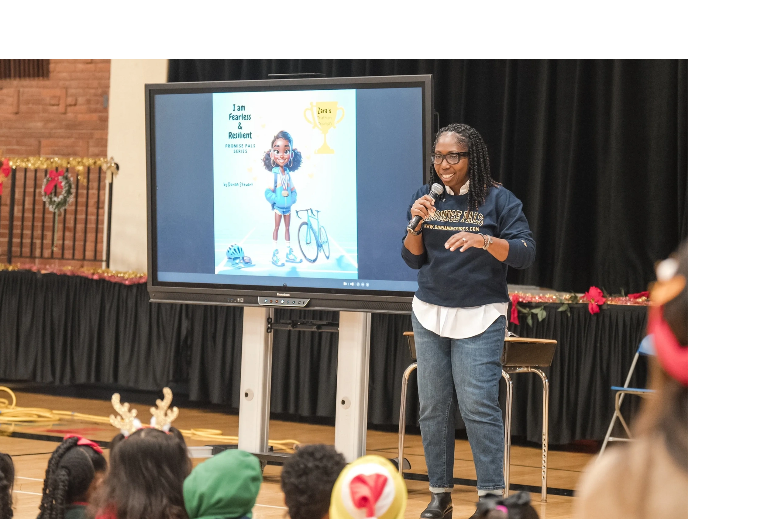 A woman speaks to children with a presentation screen showing a character and motivational message.