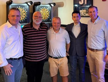 Five men posing in a room with dartboards in the background.