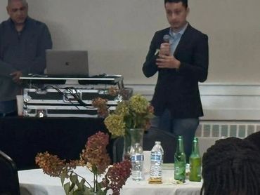 People seated at round tables listening to speakers in a conference room.