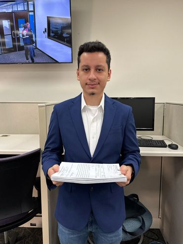 A man in a blue blazer holding a stack of papers in an office cubicle.
