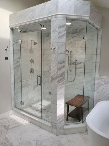 Modern glass-enclosed dual shower with marble tiles and a wooden bench inside.