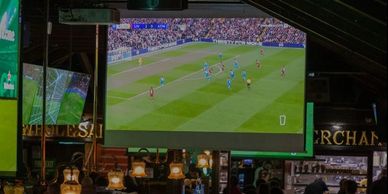 Indoor bar with large screen showing a soccer match.
