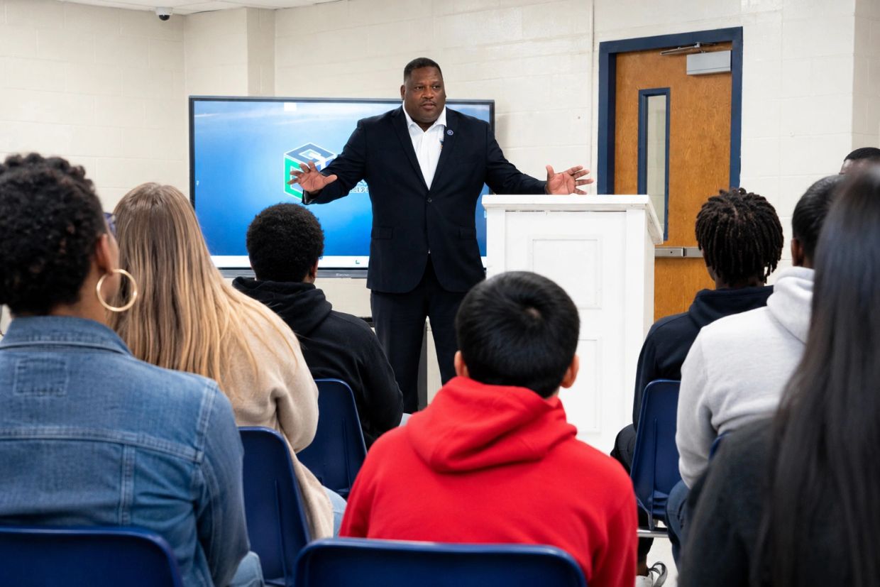 Man in suit speaking to an attentive audience in a classroom.