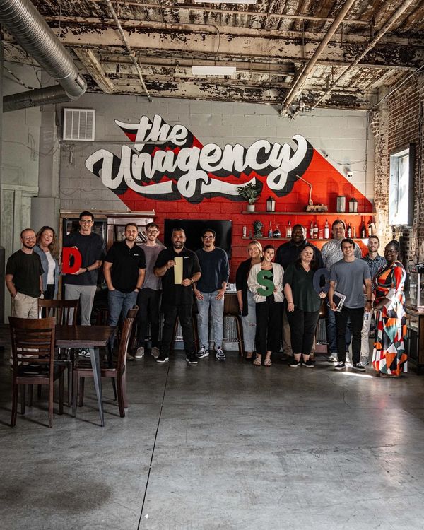 Diverse group of people posing in front of a mural with the text 'The Unagency'.