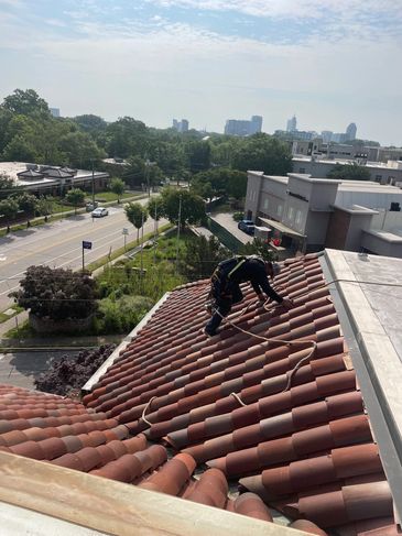 A worker inspects a tiled roof secured with safety ropes overlooking a cityscape.