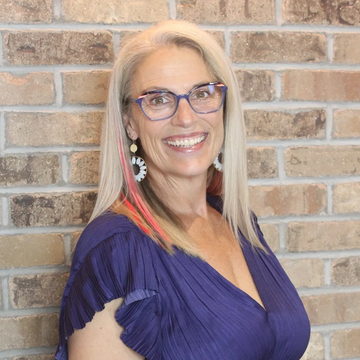 Smiling woman with glasses and colorful hair streaks in front of a brick wall.