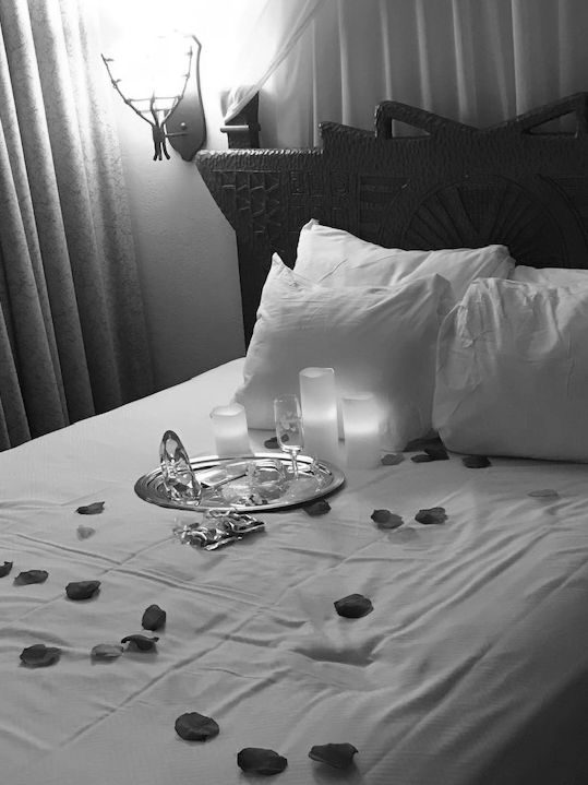 bed with  white blanket sprinkled with red rose petals  light shines on drapes behind the headboard
