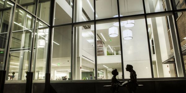 Photo of "A World to Teach" statue silhouetted against lit up interior of Ames Public Library