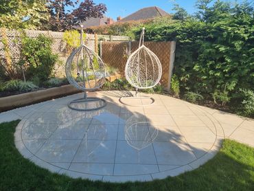Two hanging wicker chairs on a tiled circular patio in a sunny garden.
