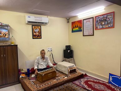 Man playing harmonium in a decorated room with religious paintings and a speaker.