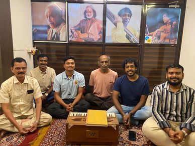 Group of six men sitting on a carpet with a harmonium, portraits of musicians behind.