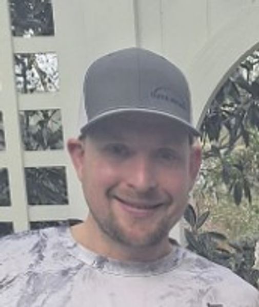 Smiling man wearing a gray cap and patterned shirt outdoors.