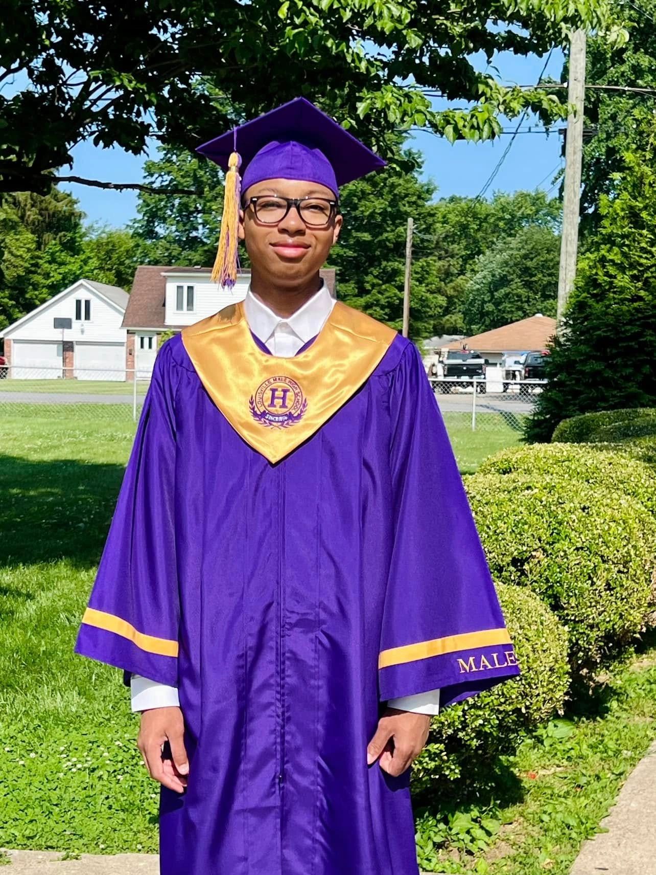 A graduate in a purple and gold cap and gown standing outdoors.
