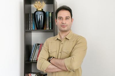 Man in beige shirt with arms crossed standing beside a bookshelf.