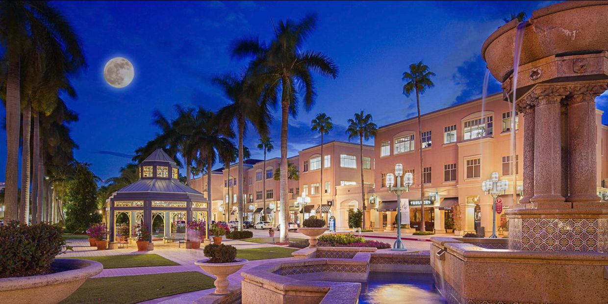 Nighttime scene of a lit plaza with palm trees and a fountain under a full moon.