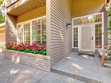 Entry door in light tan/brown color surrounded by similar color siding and red flowers.