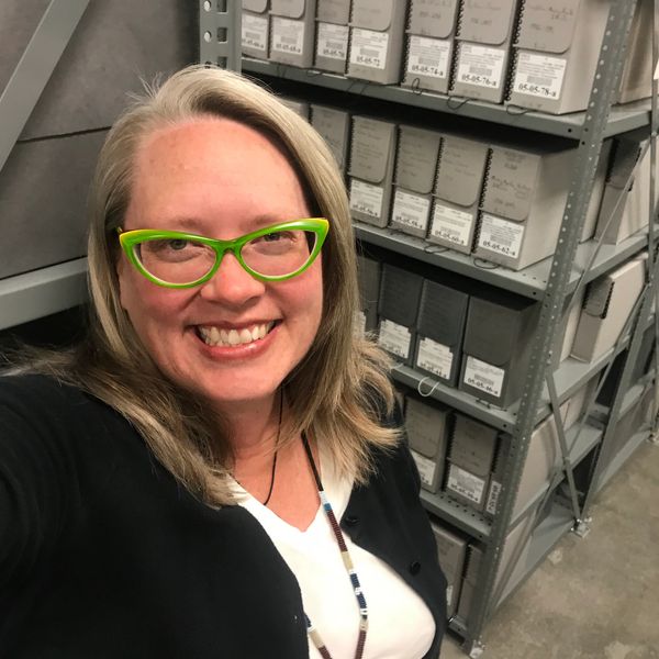 Mary B Hansen wearing green glasses and a black cardigan, standing in front of gray archival boxes.