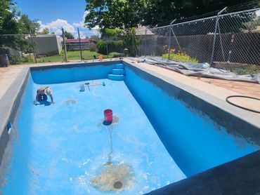 Man working on resurfacing an empty backyard swimming pool under a sunny sky.