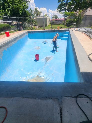 A person cleaning and painting an empty backyard pool on a sunny day.