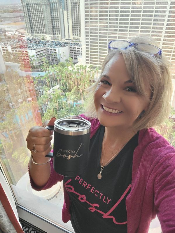 Smiling woman holding a "Perfectly Posh" mug by a window with city view.