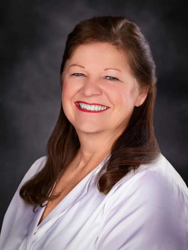 Smiling mature woman in white blouse with dark hair against a dark background.