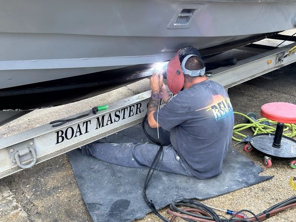 A man welds the underside of a boat on a trailer labeled 'BOAT MASTER'.