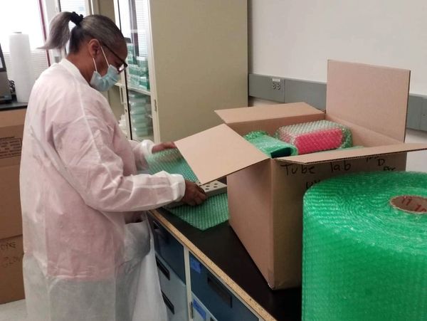 A woman in a lab coat and mask packing an open cardboard box with bubble wrap