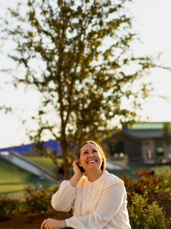 A woman in a white blouse smiles joyfully outdoors under soft sunlight.
