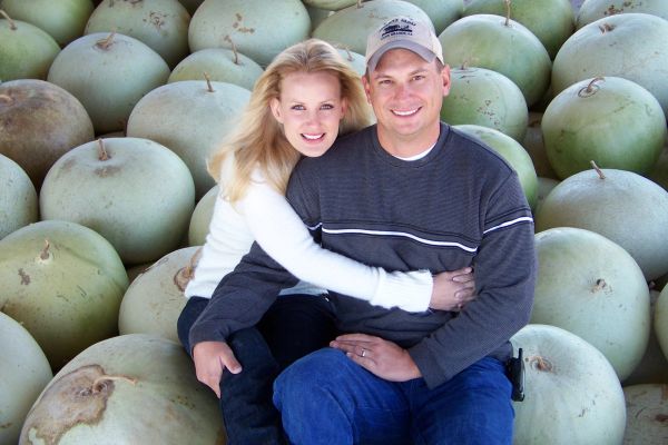 Smiling couple sitting among large melons, embracing happily.