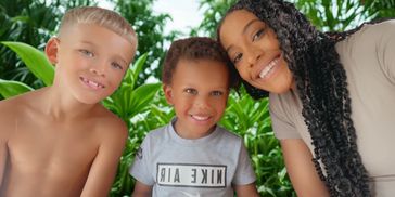 A woman and two boys smiling outdoors with green plants in the background.