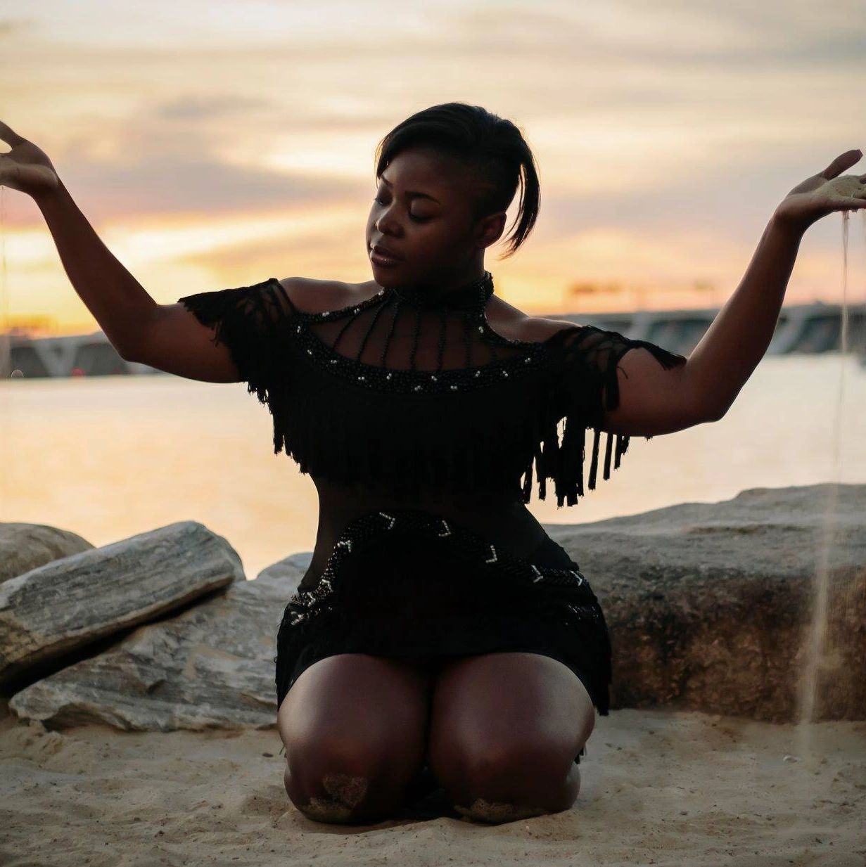 Woman in black dress kneeling on sand with arms raised at sunset.