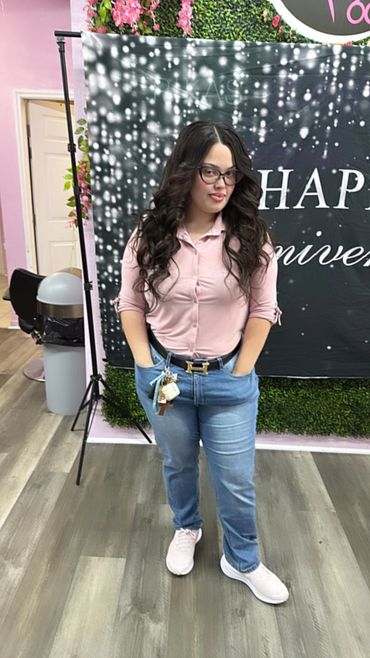 Woman with long curly hair wearing glasses, pink shirt, and jeans posing indoors.