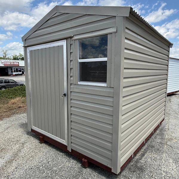 Small beige storage shed with a single door and window on gravel ground.