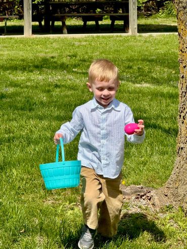 A young boy joyfully holding a blue basket and a pink Easter egg outdoors.