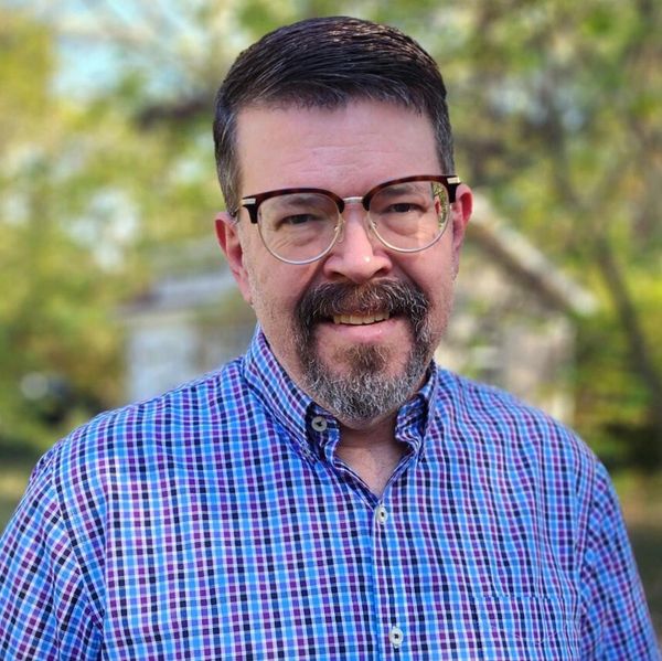 Smiling man with glasses and a checkered shirt outdoors.