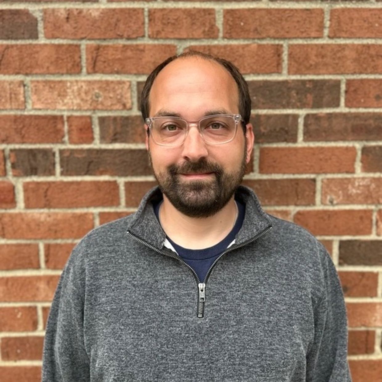 Man with glasses and beard smiling against a brick wall.