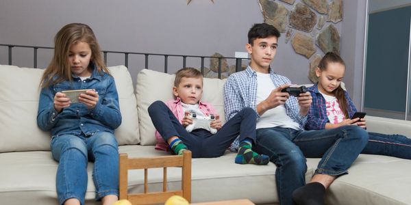 Four kids sitting on a couch, each playing on handheld gaming devices.