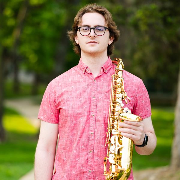 Young man in a pink shirt holding a saxophone outdoors.
