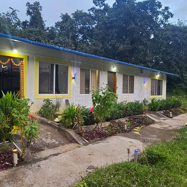 A long single-story building with yellow and white walls, surrounded by greenery and a pathway.