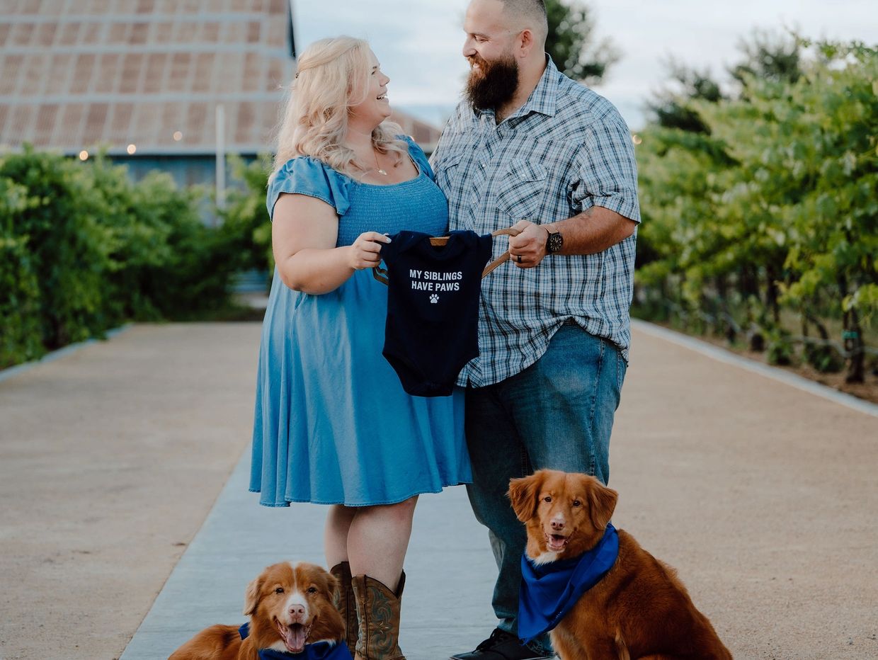 Couple holding baby onesie with two dogs.