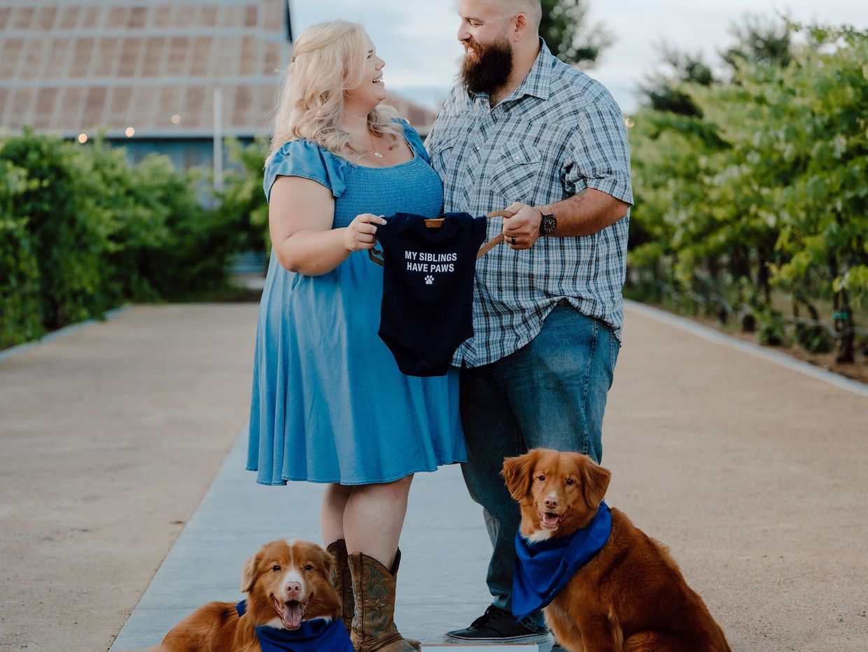 Couple holding baby onesie with two dogs.