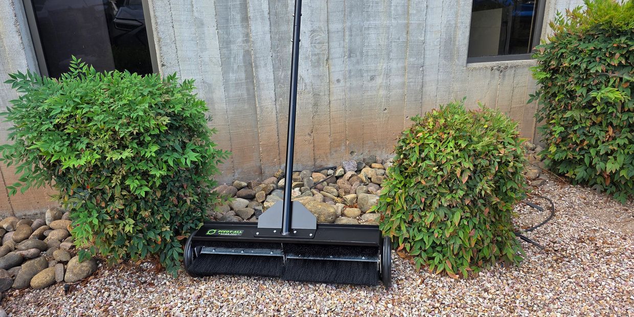 A black outdoor electric sweeper between green bushes on gravel.
