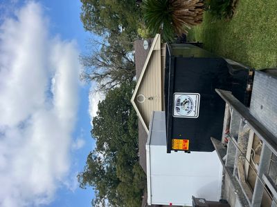 A black dumpster and trailer in a residential backyard with trees and a small house.