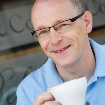 Image of Jonathan Morris raising a coffee cup outside a cafe. 