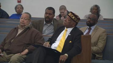 Men and women seated in a church pew, one man in a black suit with a yellow tie taking notes.